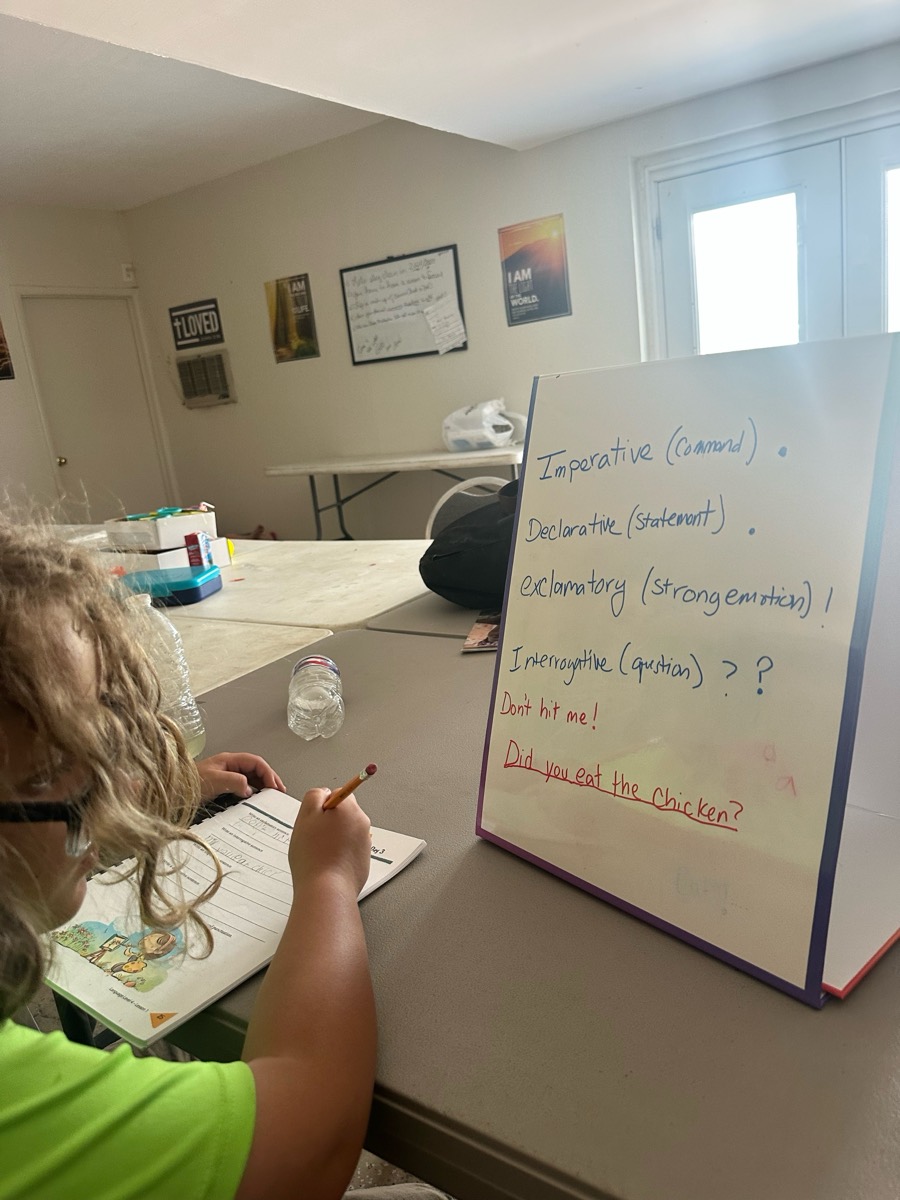 A student working in a notebook next to a whiteboard grammar lesson on the four kinds of sentences